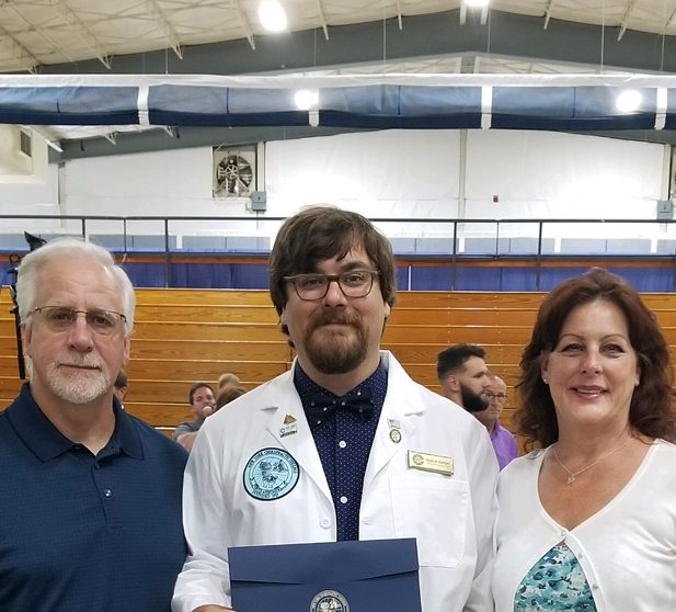 Human doctor of chiropractic, Ryan Kiehart, wearing dark blue button up with blue tie and white coat.  Flanked by humans Roger and Christine Kiehart. Photo taken in NYCC gymnasium. 
