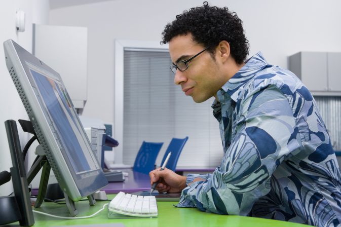 Image of human with poor computer posture. He is seated at a green desk with a white keyboard and silver monitor. He wears multicolored blue shirt and black glasses. 